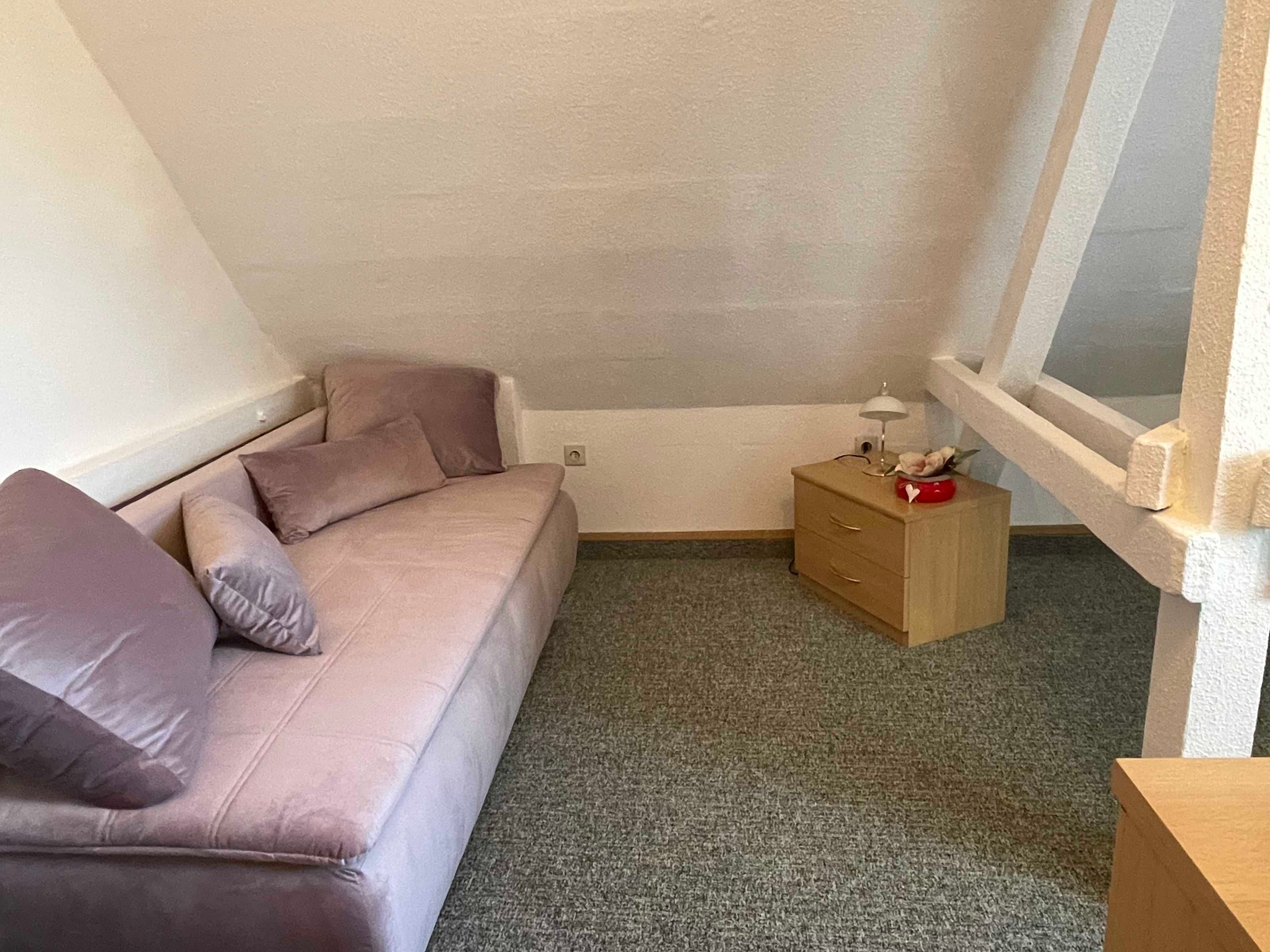 A small, cozy room with a single sofa, a wooden nightstand, and a window.