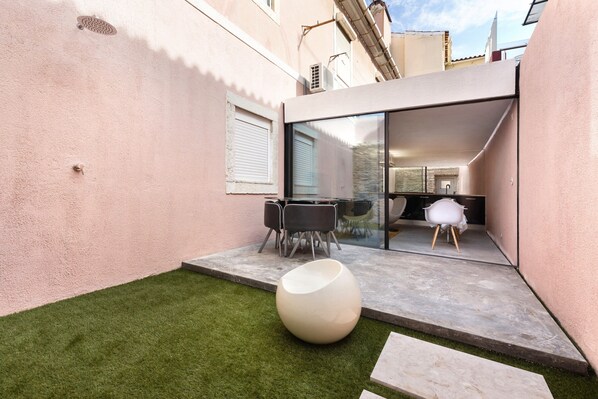 A modern outdoor patio with a glass door leading to a kitchen, a round white seating area, and a small grassy patch.