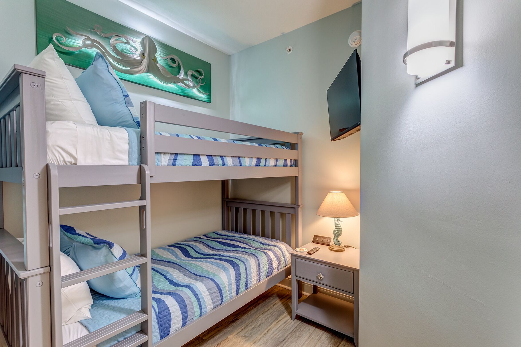 Une chambre avec un lit superposé, une table de chevet et une lampe, un tableau au mur et un luminaire au plafond.