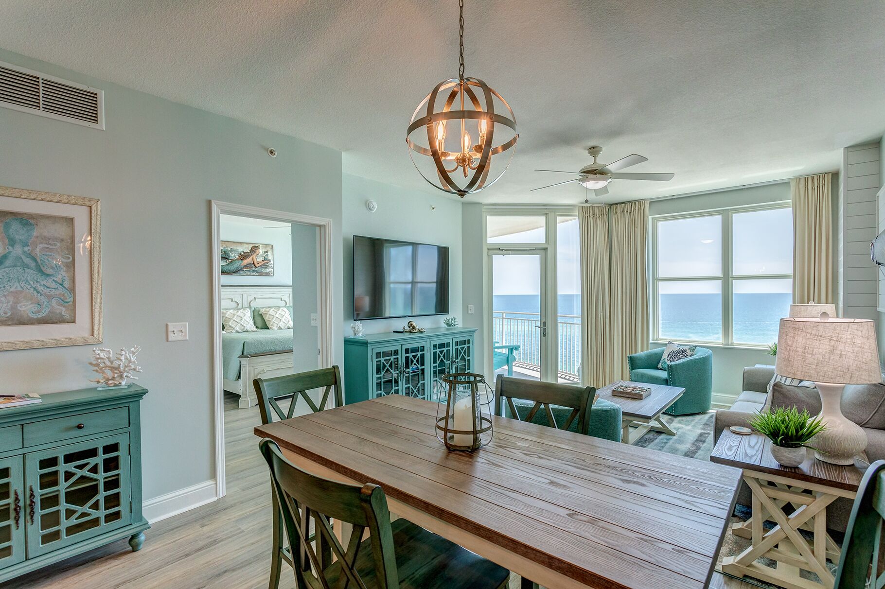 A spacious living room with a wooden dining table, teal furniture, a large TV, and a balcony with ocean views.