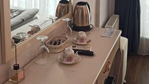 Habitación de hotel con escritorio, televisor montado encima, espejo, tetera, cafetera y ventana con cortinas.