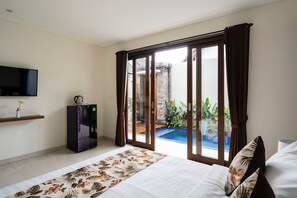 A hotel room with a bed, a flat-screen TV, a minibar, and a view of a swimming pool. - The Calna Villa (Kuta)