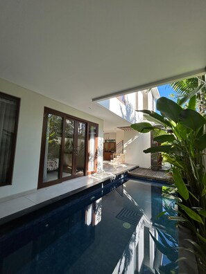 A modern indoor swimming pool with clear glass walls, a wooden deck, and a view of the surrounding greenery. - The Calna Villa (Kuta)
