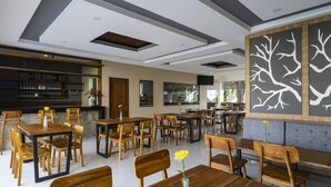 A spacious restaurant with wooden tables and chairs, a bar area, and a decorative wall with tree designs. - The Calna Villa (Kuta)