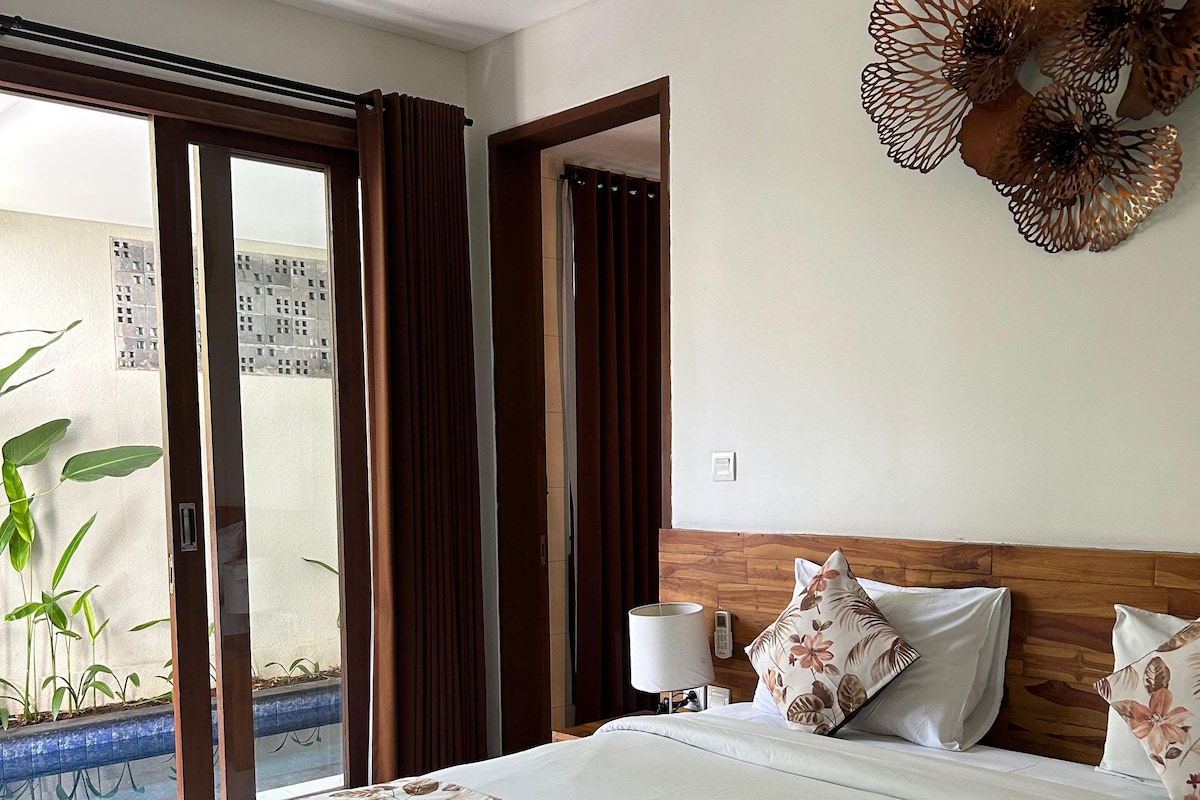 Habitación de hotel con una cama grande, cabecera de madera, mesita de noche con lámpara y vista a una piscina a través de una puerta corrediza.