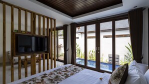 A bedroom with a large bed, a TV mounted on a wooden partition, and a view of an outdoor swimming pool. - The Calna Villa (Kuta)