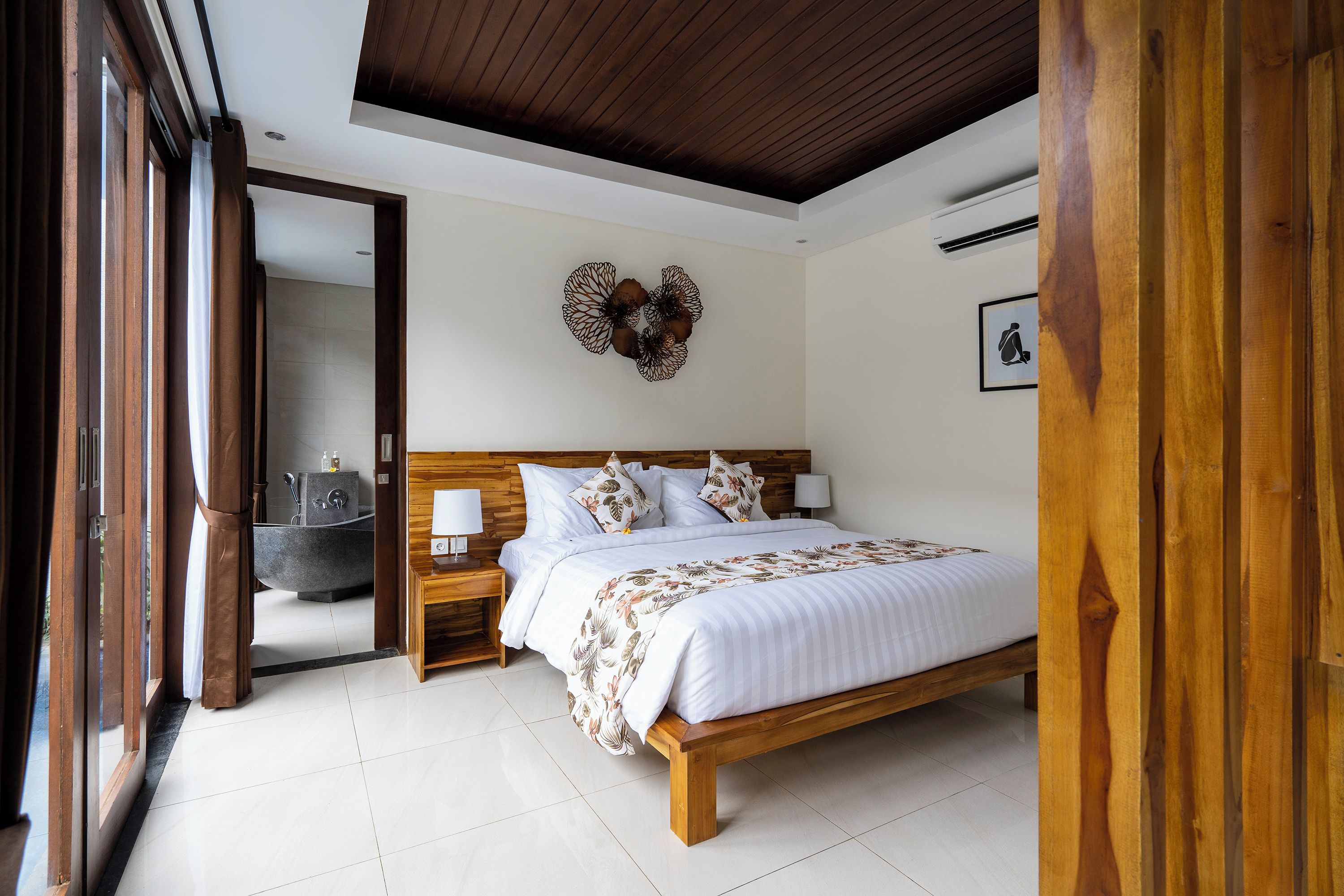 Une chambre à coucher avec un lit en bois, une table de chevet, une lampe fixée au mur, et une salle de bain visible à travers une porte ouverte.