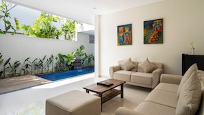 A modern living room with a beige sofa, a wooden coffee table, and a small side table. There is a large window with a view of a swimming pool and greenery outside. - The Calna Villa (Kuta)