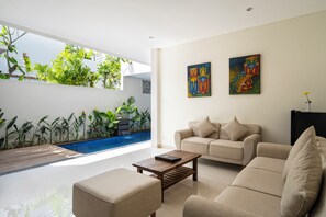 A modern living room with a beige sofa, a wooden coffee table, and a small side table. There is a large window with a view of a swimming pool and greenery outside. - The Calna Villa (Kuta)