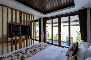A bedroom with a large bed, a TV mounted on a wooden partition, and a view of an outdoor swimming pool. - The Calna Villa (Kuta)