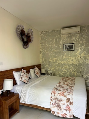 A bedroom with a wooden headboard, a bed with white bedding and a floral-patterned throw, a nightstand with a lamp, and a wall-mounted air conditioner. - The Calna Villa (Kuta)