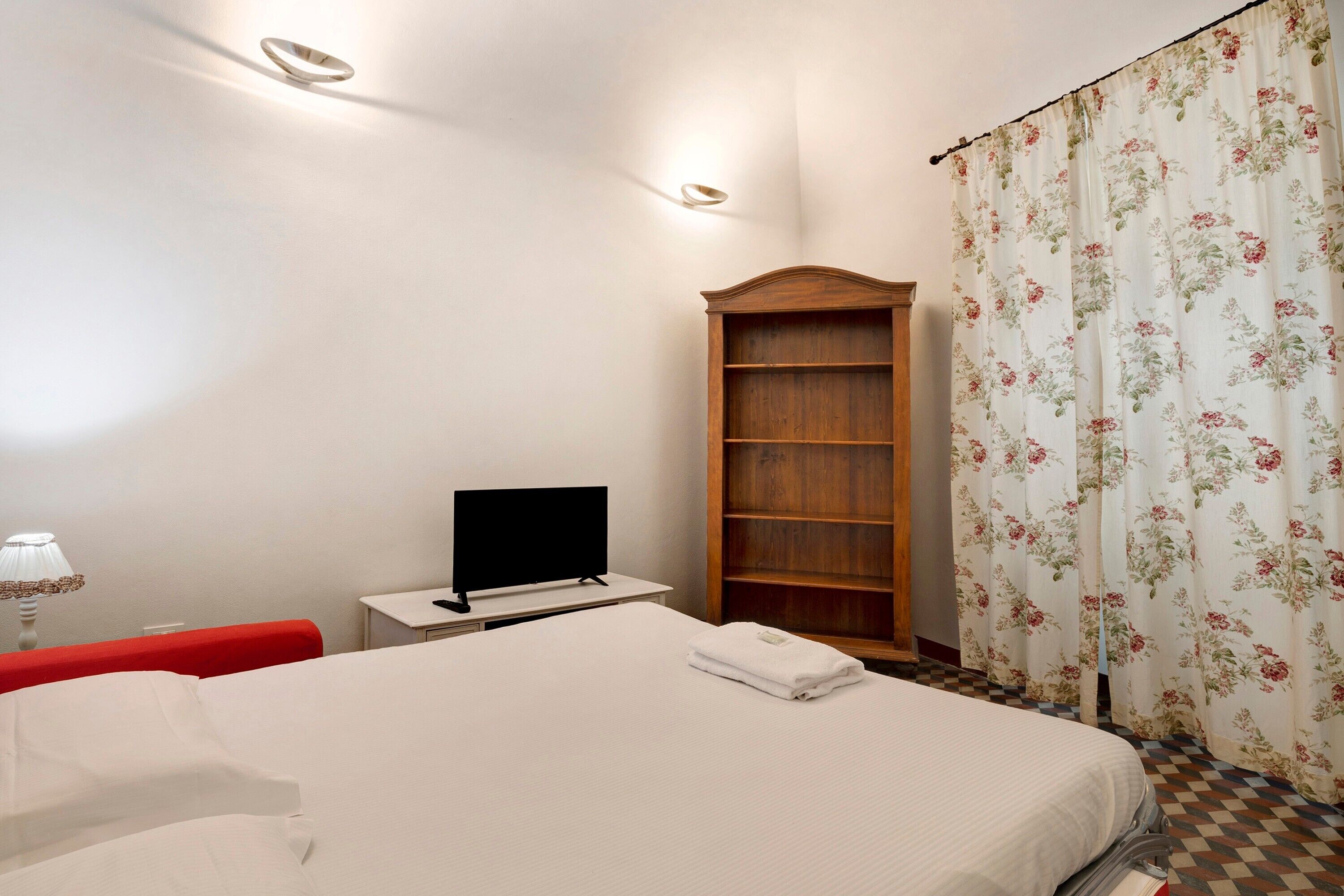 Ett hotellrum med en säng, en tv på ett litet bord, en bokhylla i trä och ett fönster med blommiga gardiner.