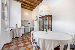 Una cucina con pavimento a scacchi, mobili bianchi, un tavolo da pranzo con tovaglia bianca e una lampada a sospensione.