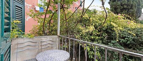 Un balcón con una mesa redonda y sillas, barandilla de metal y vista a un edificio rosa y follaje.
