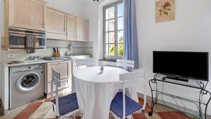 A compact kitchen with a washing machine, a dining table with chairs, a TV on a stand, and a window with curtains.
