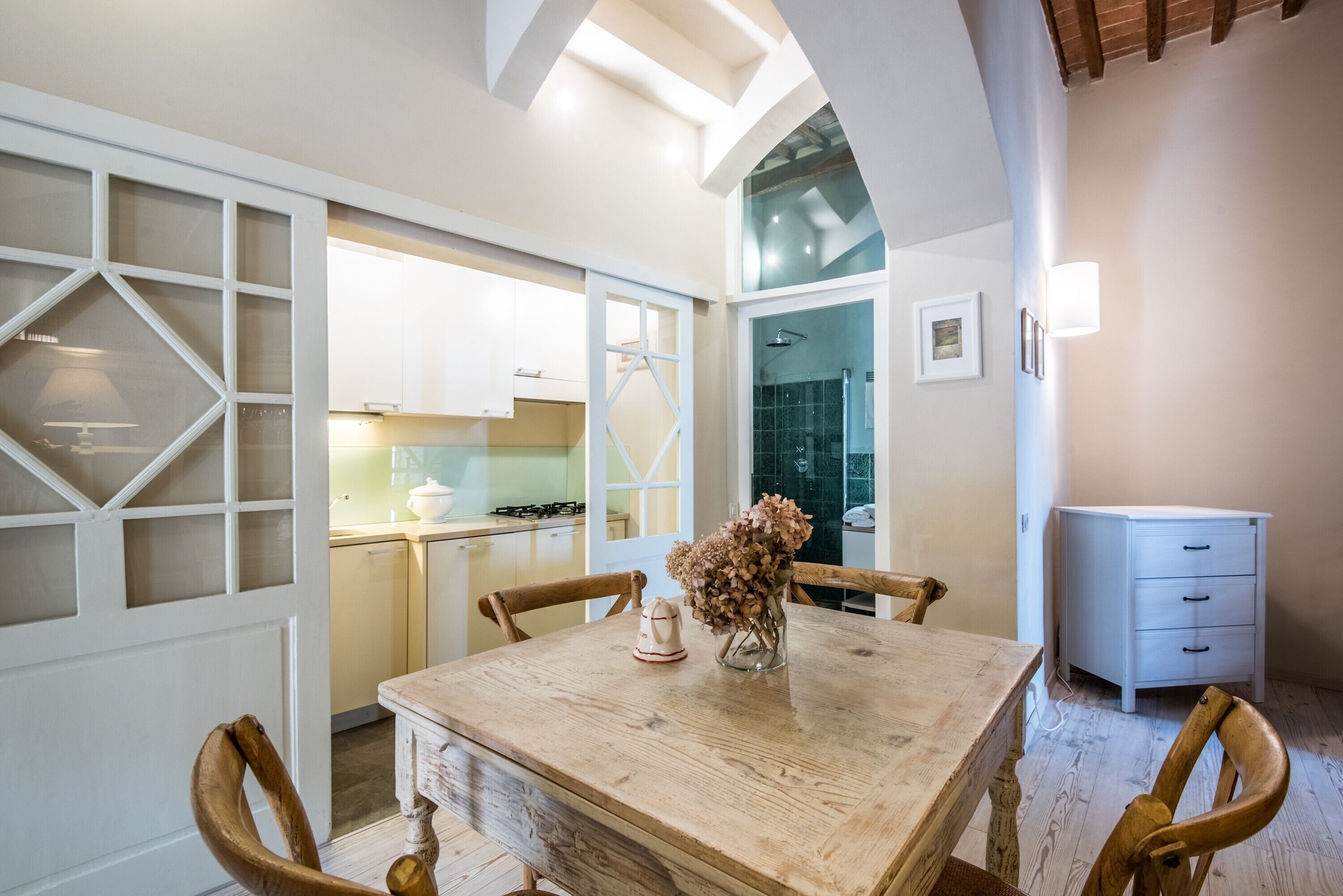 A dining area with a wooden table, chairs, a vase with dried flowers, and a view of a kitchen and bathroom through an open door.