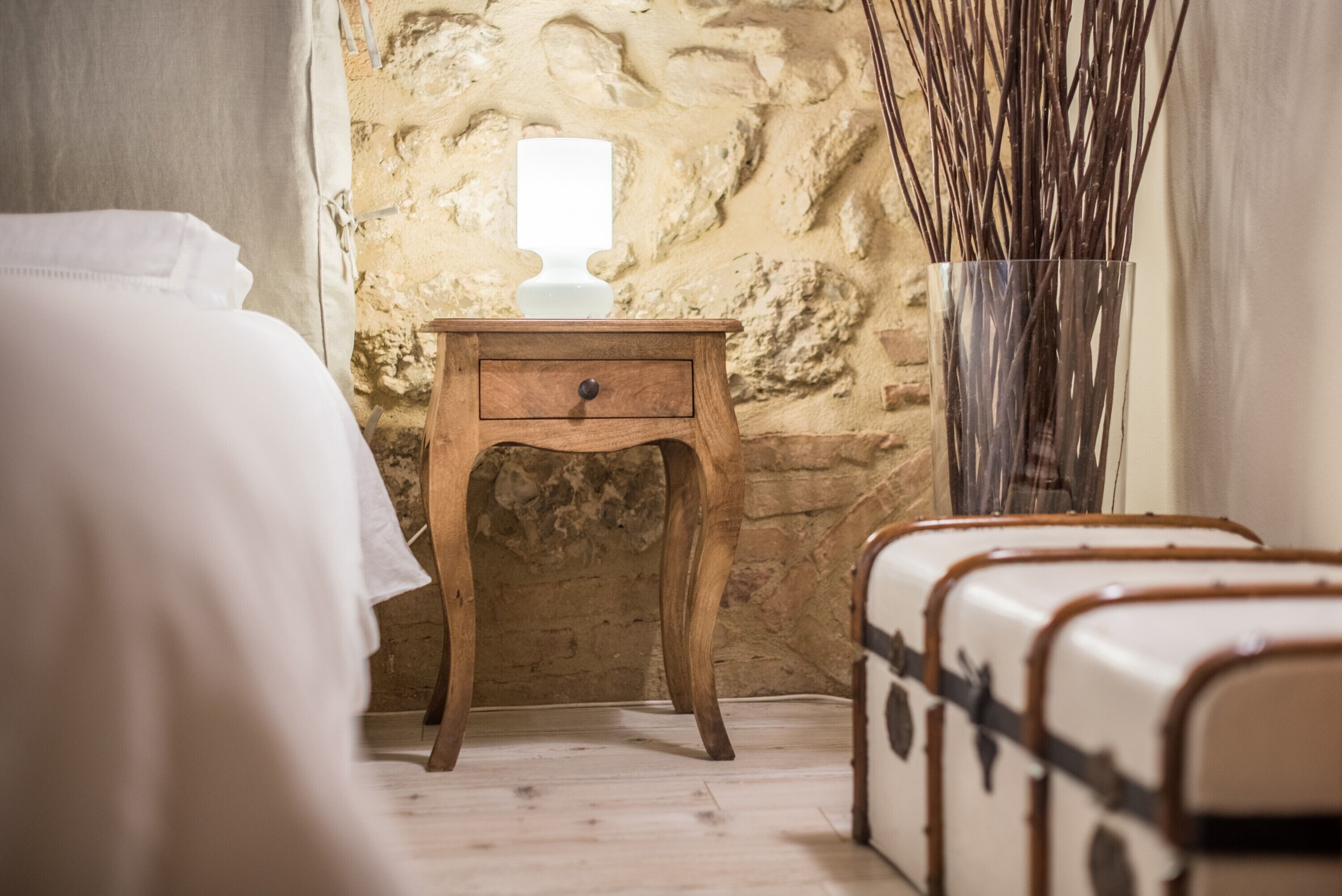 Een kamer met een bed, een houten nachtkastje met een lamp, een stenen muur en oude koffers.