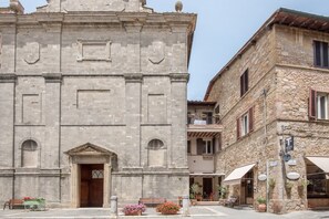 Un edificio histórico de piedra con una entrada central, un balcón en el piso superior y un pequeño espacio al aire libre con mesas y sillas.