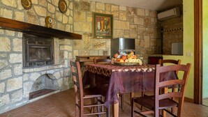 Une pièce aux murs de pierre, avec une cheminée, une table à manger entourée de chaises et un panier de fruits.