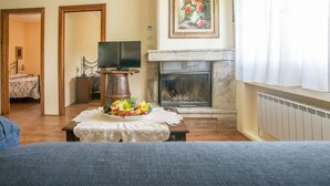 A living room with a fireplace, a television, a dining table with a fruit centerpiece, and a painting on the wall.