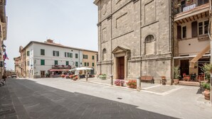 Un bâtiment historique en pierre, doté d’une entrée centrale, situé à côté d’une rue où se trouvent des terrasses et des plantes en pot.