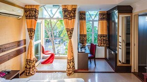 A hotel room with a bed, a desk, a chair, and a TV. There is a window with curtains and a view of greenery outside. - RIJ HULHUMALE (Hulhumalé)