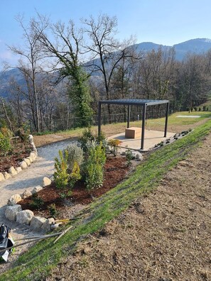 A covered outdoor seating area with a stone pathway leading to it, surrounded by landscaped greenery and trees.