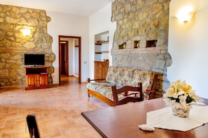 Una habitación con pared de piedra, un televisor sobre un soporte de madera, un sofá floral, una mesa de comedor y un florero con flores.