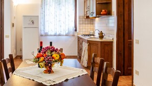 A dining table with a fruit centerpiece, wooden chairs, a kitchen with wooden cabinets, and a window with curtains.