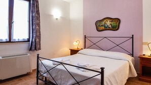 A bedroom with a bed, a radiator, a window with curtains, a nightstand with a lamp, and a wall-mounted picture.