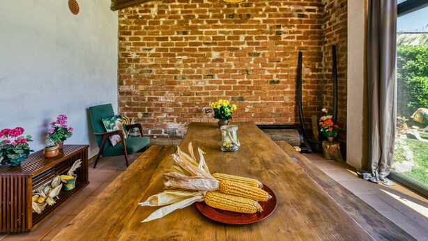 Comedor rústico con una mesa de madera, pared de ladrillo y vista a plantas verdes en el exterior.