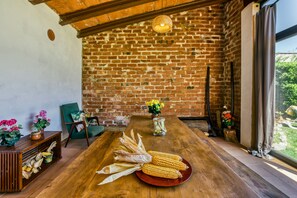A rustic dining area with a wooden table, a brick wall, and a view of greenery outside.