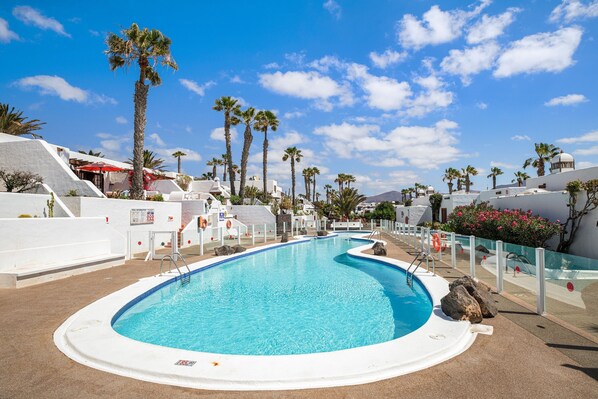 Une piscine bordée de palmiers, des bâtiments blancs et un ciel d’un bleu limpide.