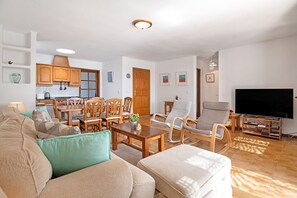 Un salon avec un canapé, des fauteuils, une table basse, un téléviseur et, au fond, une cuisine.