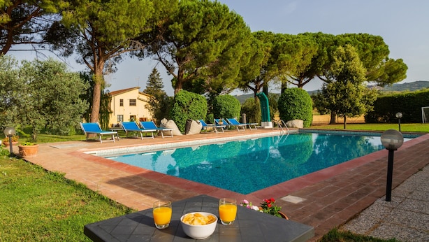 Une piscine avec des chaises longues, une table sur laquelle se trouvent un saladier et des boissons, et un bâtiment en arrière-plan.