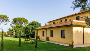 Un bâtiment jaune avec un toit recouvert de tuiles, un chemin pavé et une pelouse bien entretenue agrémentée d’arbres et de plantes en pot.
