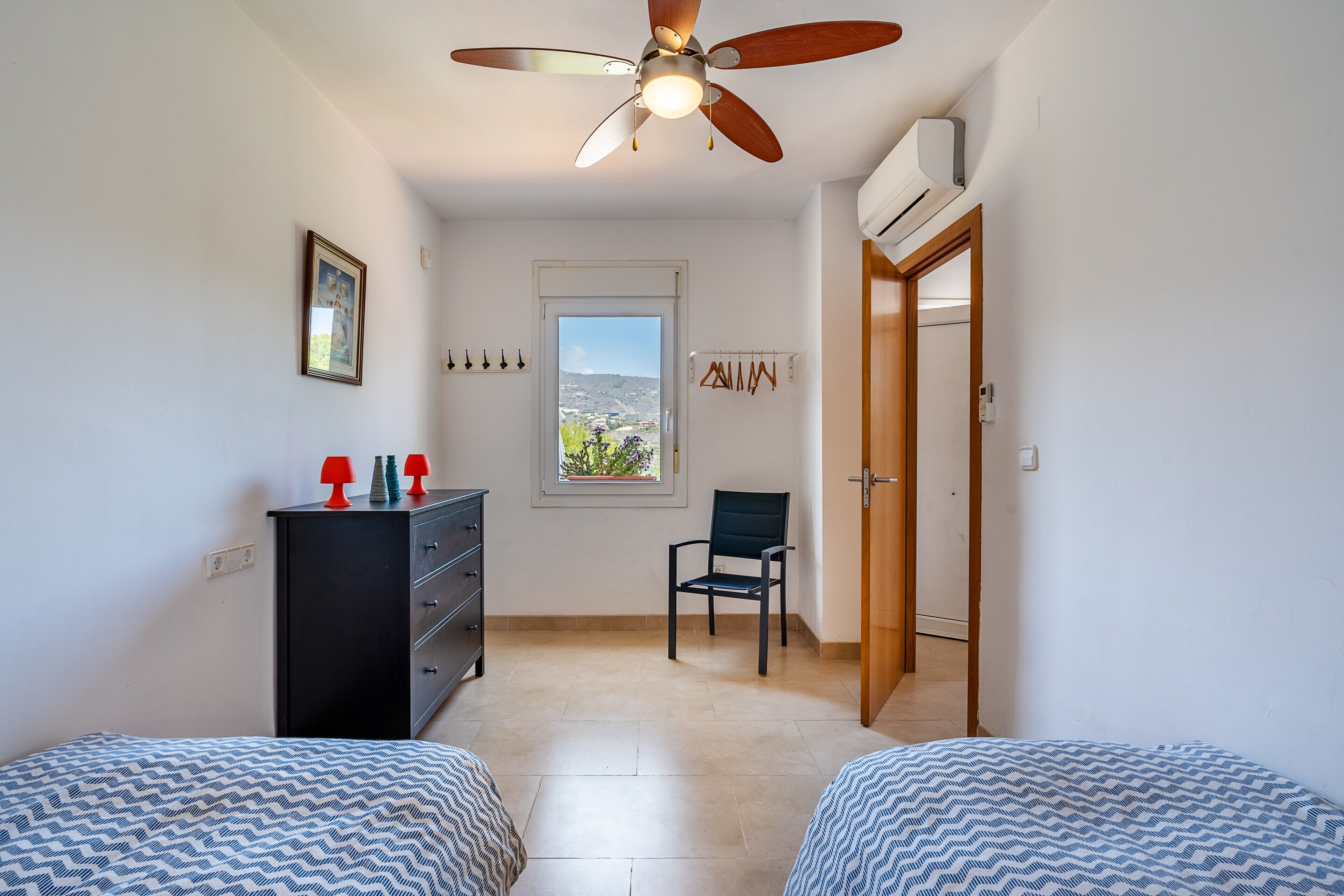 Un dormitorio con dos camas, un ventilador de techo, un aparador, una silla, una ventana con vistas y una puerta.