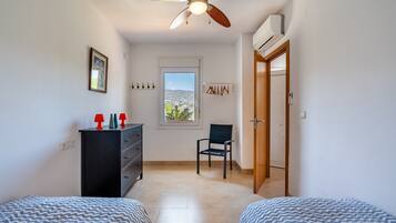 Un dormitorio con dos camas, un ventilador de techo, un aparador, una silla, una ventana con vistas y una puerta.