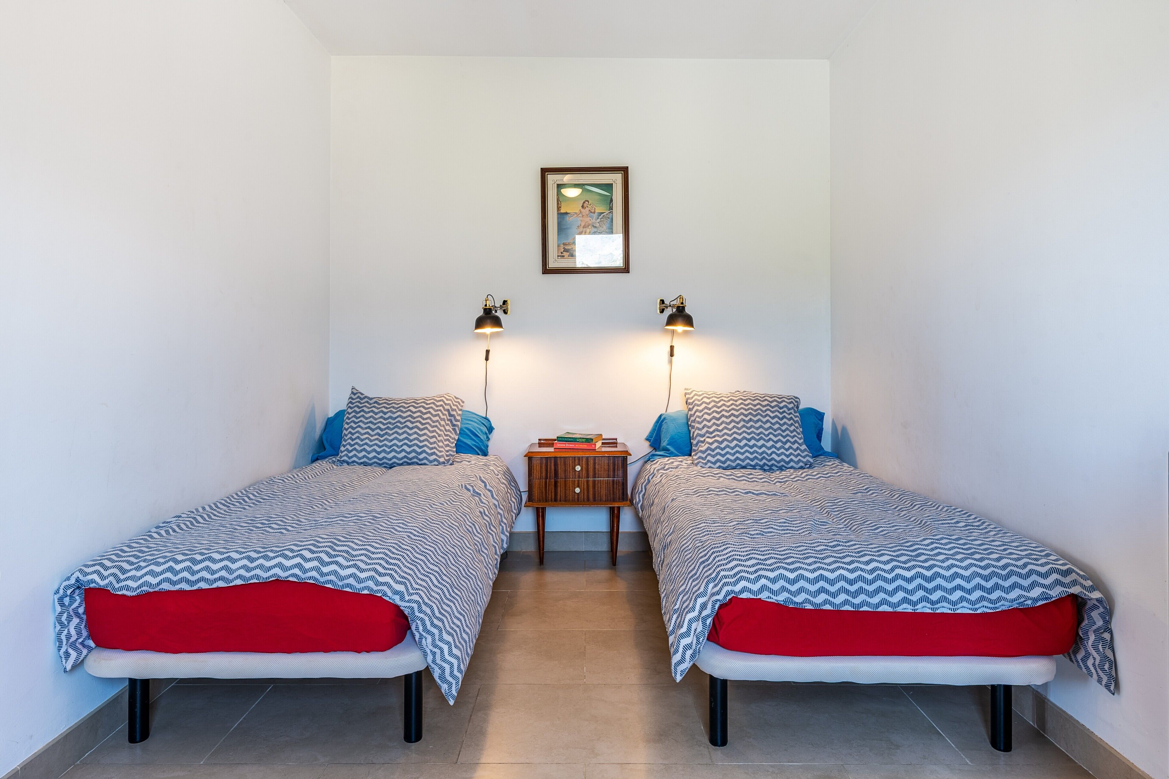 Ein Doppelzimmer mit zwei Betten, einem Nachttisch, zwei Wandlampen und einem gerahmten Bild an der Wand.
