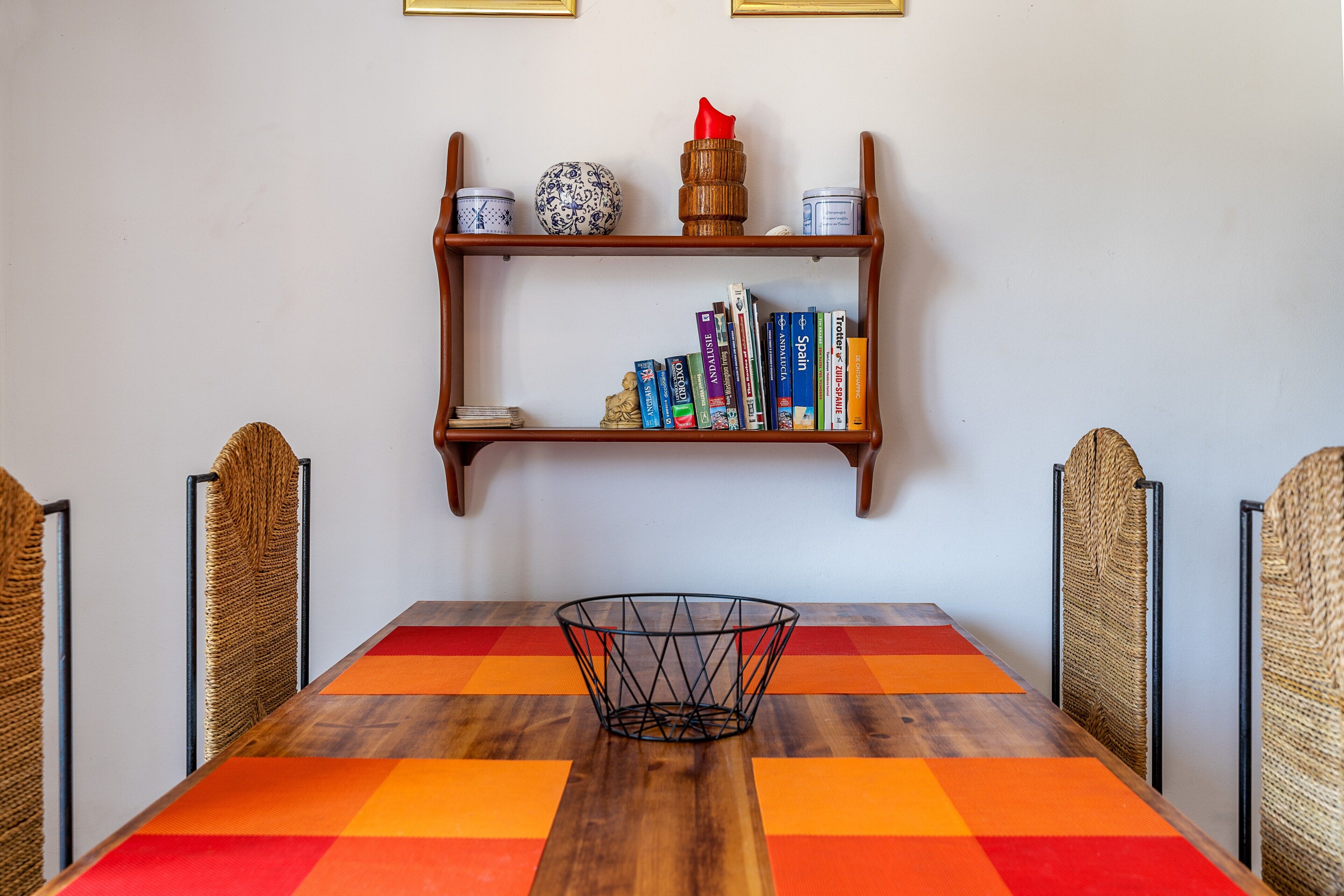 Une table à manger agrémentée d’un chemin de table aux couleurs vives, un panier en métal placé au centre, et une bibliothèque avec des livres et des objets décoratifs au-dessus.