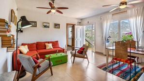 A spacious living room with a red sofa, wooden chairs, a green trunk, and a dining table with chairs. There are ceiling fans, a bookshelf, and a view of the outdoors.
