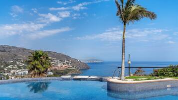 Piscina infinita con vista al mar, palmeras y un paisaje montañoso.