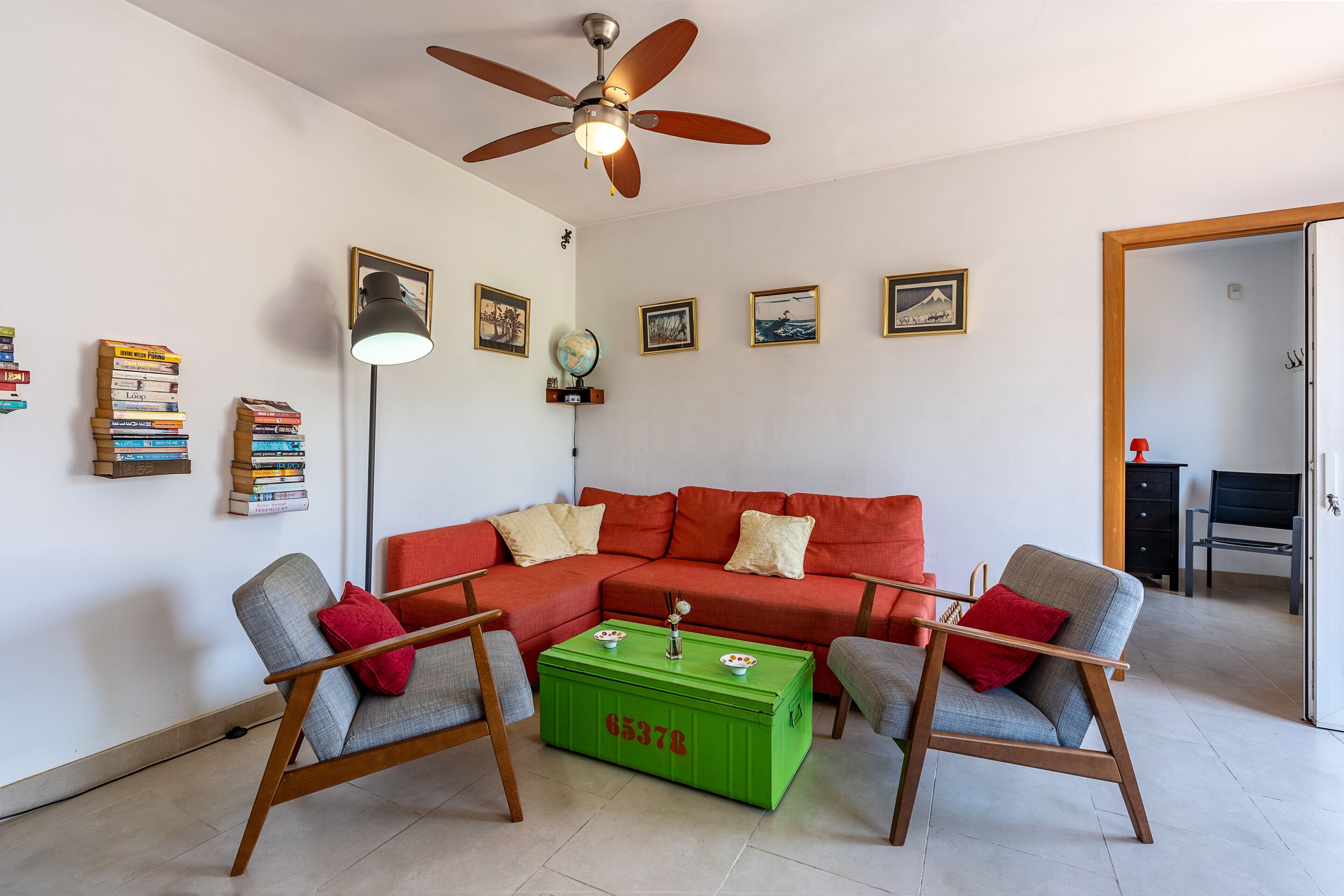 Una sala de estar con un sofá rojo, dos sillones, un aparador verde, un ventilador de techo y cuadros colgados en la pared.