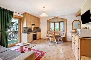 A cozy kitchen with wooden cabinets, a dining area with chairs, a large window, and a television mounted on the wall.