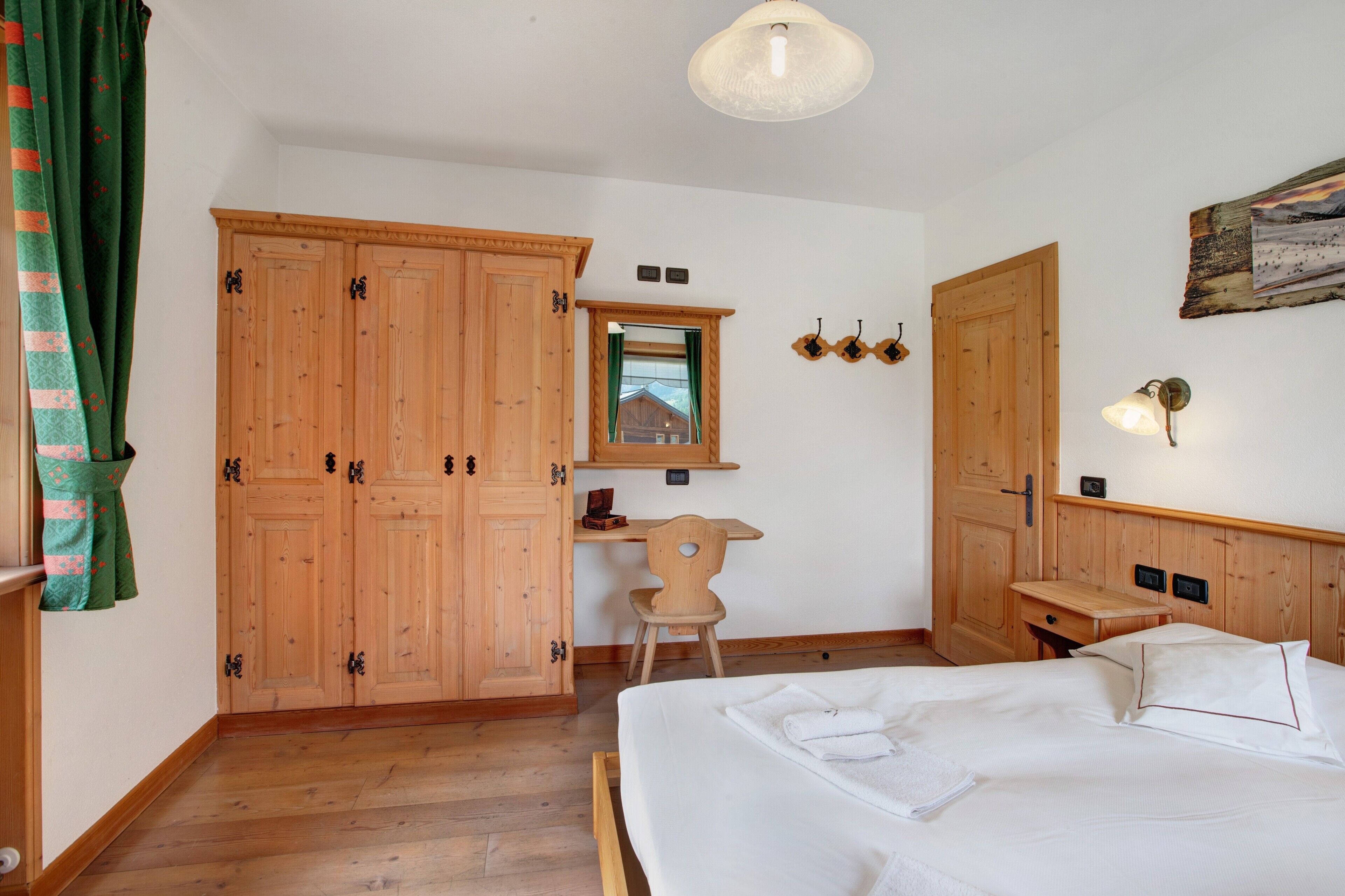 A bedroom with a wooden wardrobe, a desk with a chair, a bed with white linens, and a hanging light fixture.