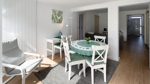 Un comedor con una mesa redonda, sillas blancas y un mantel verde. Hay una silla blanca con cojín, una lámpara y una planta sobre la mesa.