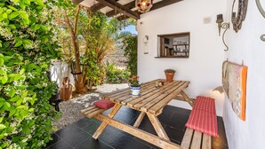 A wooden table with benches, surrounded by greenery and a white wall with a window and wall-mounted lights.