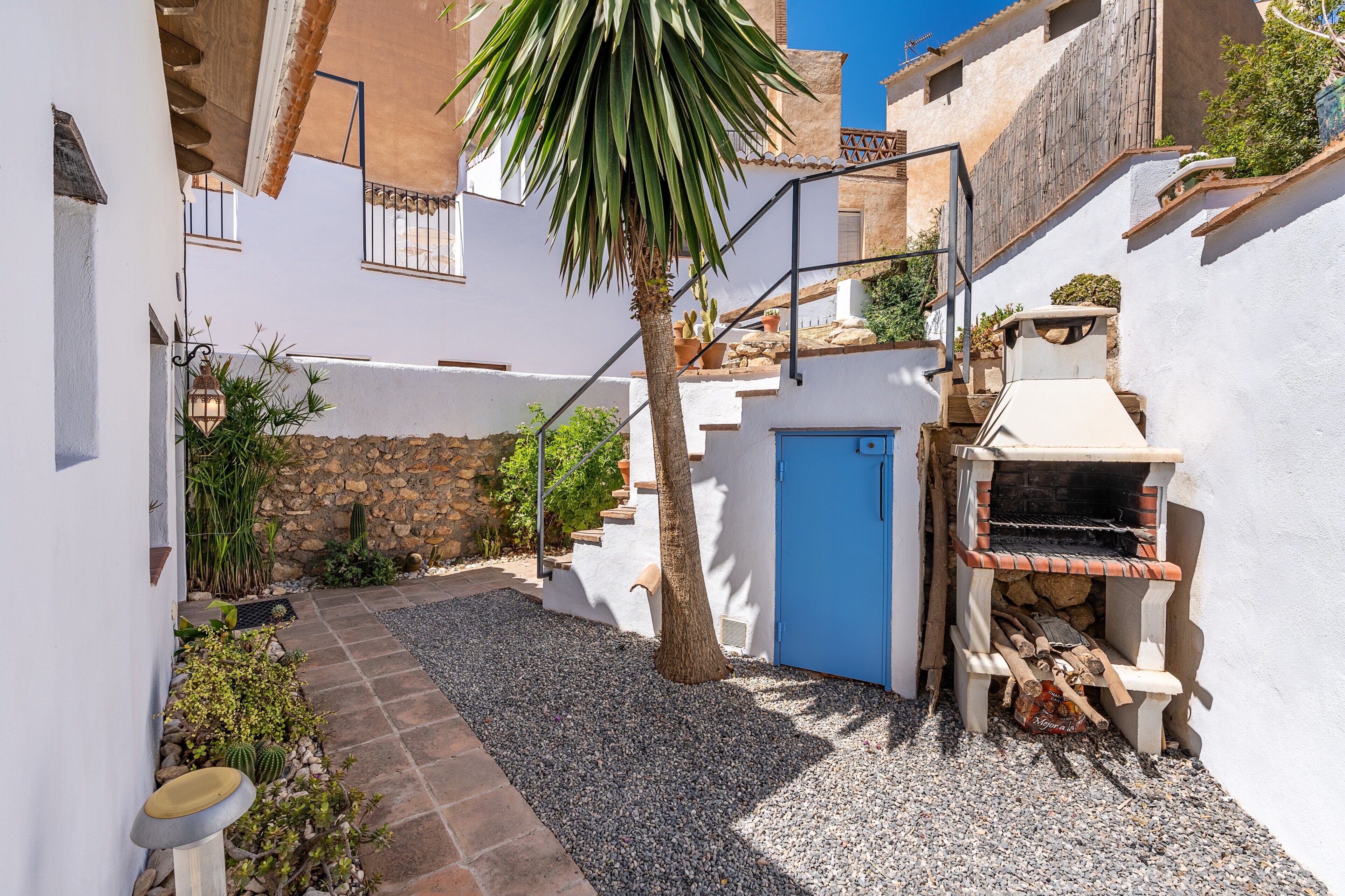 A narrow courtyard with a blue door, a stone wall, a small tree, and a wood-fired oven.