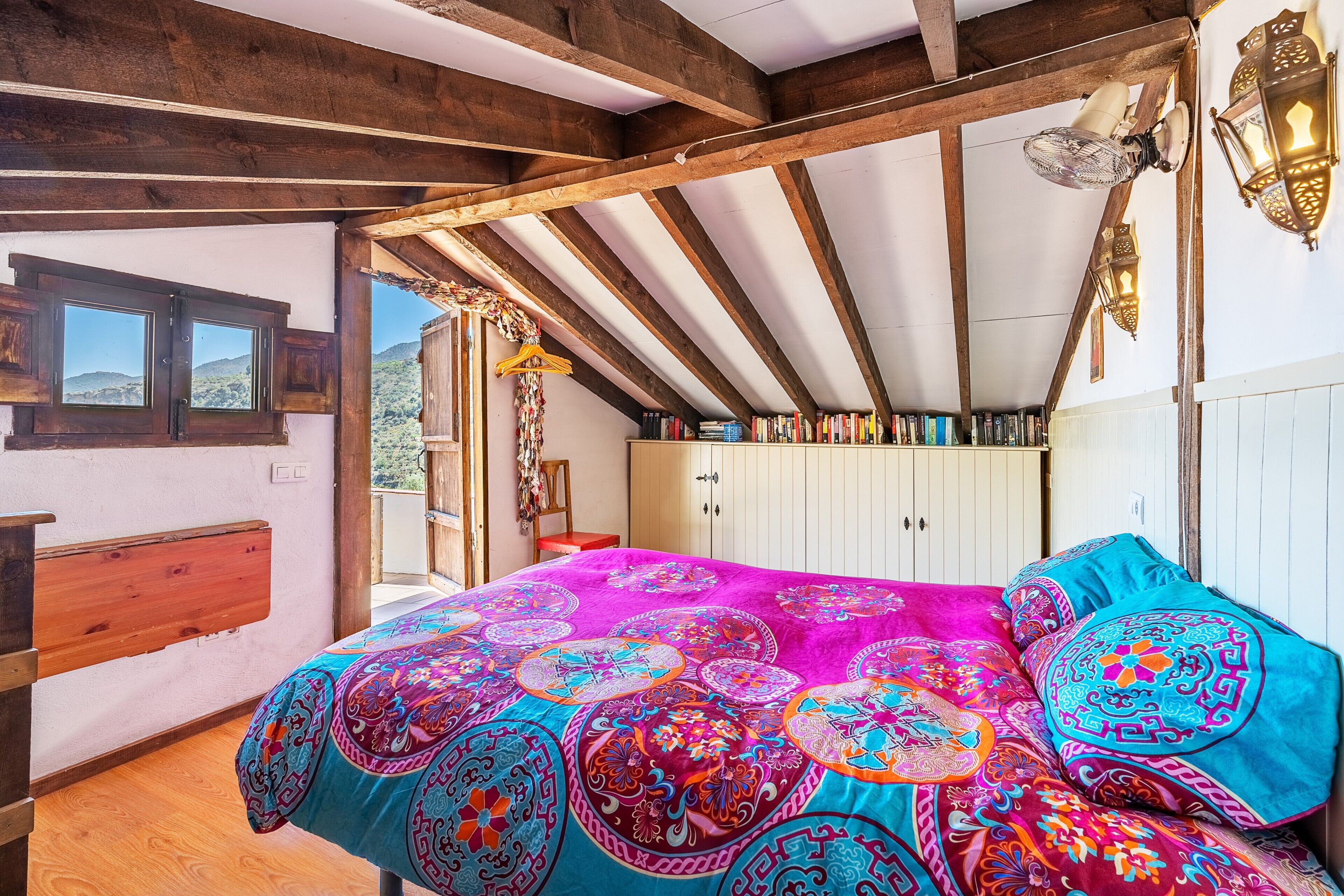 A bedroom with a colorful bedspread, wooden beams, a bookshelf, and a view through a window.