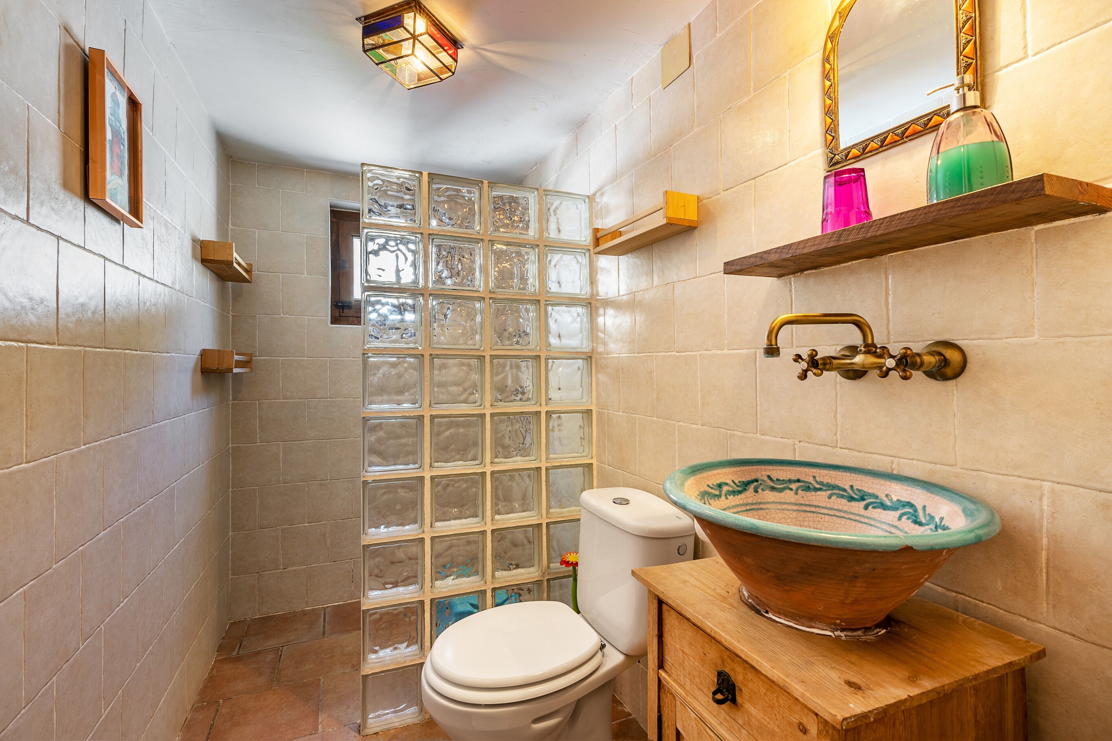 Un baño con pared de azulejos, cabina de ducha con mampara de vidrio, inodoro, tocador de madera con lavabo y espejo con marco decorativo.